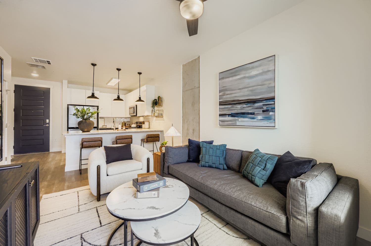 Open-concept apartment living room with a gray sofa and a view into the modern kitchen with a large island and bar seating at Avian Apartments in Colorado Springs, CO.